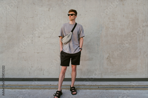 Portrait of handsome confident stylish hipster man in sunglasses with bag handy, standing guy near the concrete wall. Calm relaxing moment, summer vacation concept