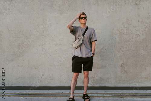 Portrait of handsome confident stylish hipster man in sunglasses with bag handy, standing guy near the concrete wall. Calm relaxing moment, summer vacation concept