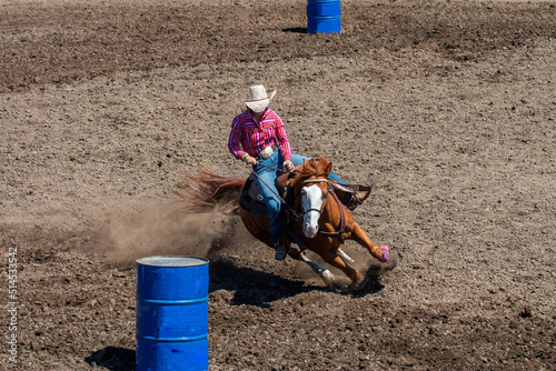 A rodeo cowgirl in a red shirt is riding a brown horse in a barrel racing competition. They are going around the barrel on the left side.  The horse is kicking up a lot of dirt.