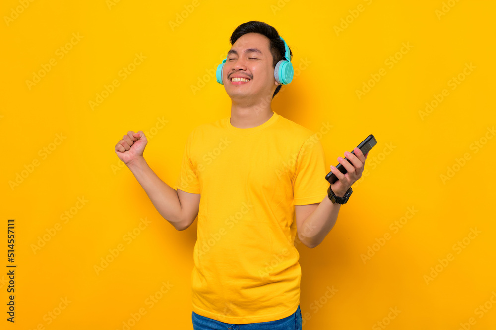 Delighted young Asian man in casual t-shirt wearing technology wireless headphones for listening to music via mobile phone isolated on yellow background. People emotions lifestyle concept Delighted young Asian man in casual t-shirt wearing technology wireless headphones for listening to music via mobile phone isolated on yellow background. People emotions lifestyle concept