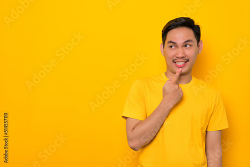 Thoughtful young Asian man in casual t-shirt looking up at copy space and touching chin, thinking about interesting offer isolated on yellow background. People lifestyle concept