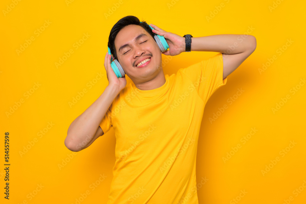 Relaxed young Asian man in casual t-shirt listening music in headphones isolated on yellow background. People emotions lifestyle concept Relaxed young Asian man in casual t-shirt listening music in headphones isolated on yellow background. People emotions lifestyle concept