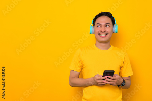 Smiling young Asian man in casual t-shirt with mobile phone and wireless headphones looking aside at copy space isolated on yellow background. People emotions lifestyle concept