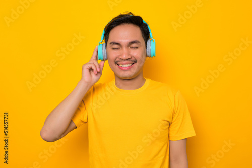 Relaxed young Asian man in casual t-shirt listening music in headphones isolated on yellow background. People emotions lifestyle concept © Sewupari Studio