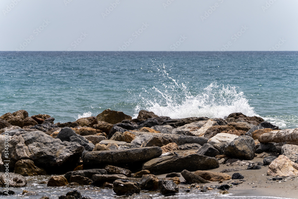 Beach on the Italian Riviera with sun, sea and stones in the sea water. Rough Sea and Foamy Waves on Shore at Mediterranean Coast in Sunrise