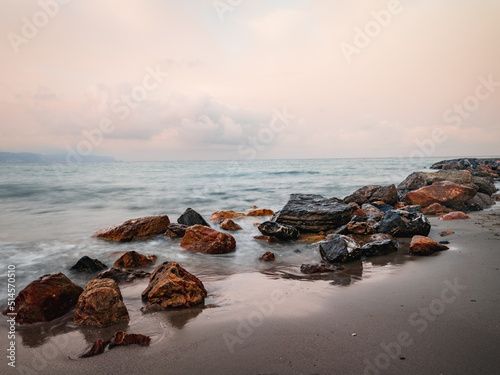 Beach on the Italian Riviera with sun, sea and stones in the sea water. Rough Sea and Foamy Waves on Shore at Mediterranean Coast in Sunrise