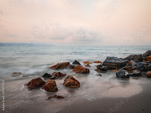 Beach on the Italian Riviera with sun, sea and stones in the sea water. Rough Sea and Foamy Waves on Shore at Mediterranean Coast in Sunrise