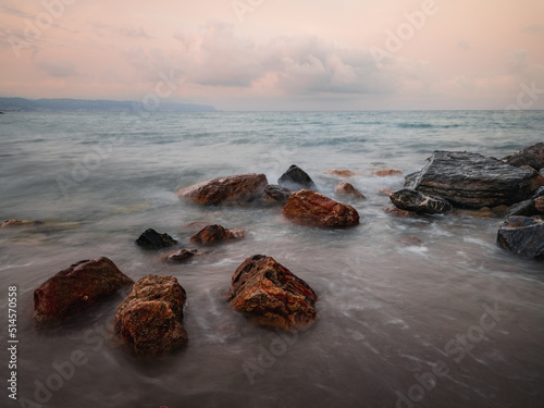 Beach on the Italian Riviera with sun, sea and stones in the sea water. Rough Sea and Foamy Waves on Shore at Mediterranean Coast in Sunrise