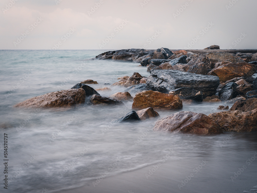 Fototapeta premium Beach on the Italian Riviera with sun, sea and stones in the sea water. Rough Sea and Foamy Waves on Shore at Mediterranean Coast in Sunrise