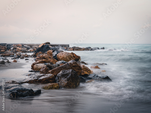 Beach on the Italian Riviera with sun, sea and stones in the sea water. Rough Sea and Foamy Waves on Shore at Mediterranean Coast in Sunrise