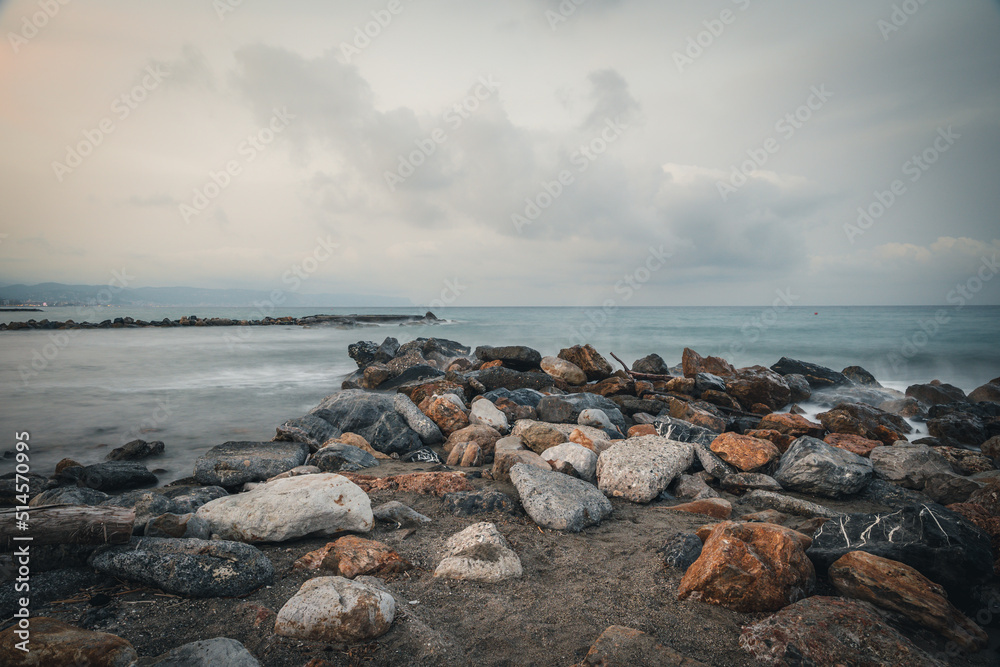 Beach on the Italian Riviera with sun, sea and stones in the sea water. Rough Sea and Foamy Waves on Shore at Mediterranean Coast in Sunrise Beach on the Italian Riviera with sun, sea and stones in the sea water. Rough Sea and Foamy Waves on Shore at Mediterranean Coast in Sunrise