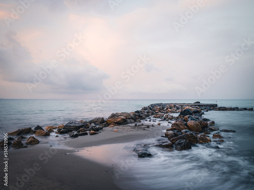 Beach on the Italian Riviera with sun, sea and stones in the sea water. Rough Sea and Foamy Waves on Shore at Mediterranean Coast in Sunrise