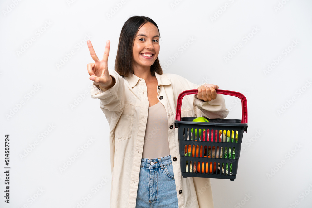 Young woman holding a shopping basket full of food isolated on white background smiling and showing victory sign Young woman holding a shopping basket full of food isolated on white background smiling and showing victory sign