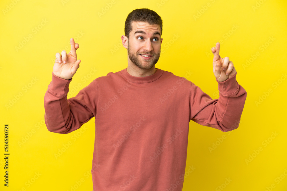 Fototapeta premium Handsome blonde man over isolated yellow background with fingers crossing and wishing the best