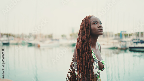 Woman with African braids wearing top looks at the yachts and ships standing on the pier in the port.