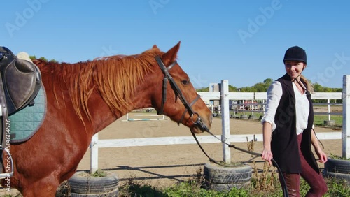 Equestrienne walks with harnessed sorrel horse in countryside. Jockette girl smiling, holding reins, looking carefully at palfrey. Young woman going to ride bay stallion on meadow in summer photo