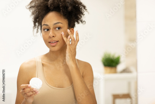 Smiling african american woman applying facial moisturizer while holding jar in bathroom
