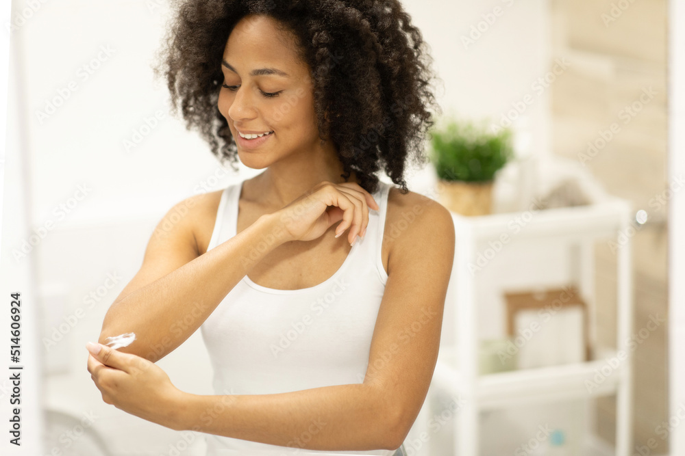 African American woman applying cream on dry elbows, moisturizing skincare while standing in bathroom African American woman applying cream on dry elbows, moisturizing skincare while standing in bathroom