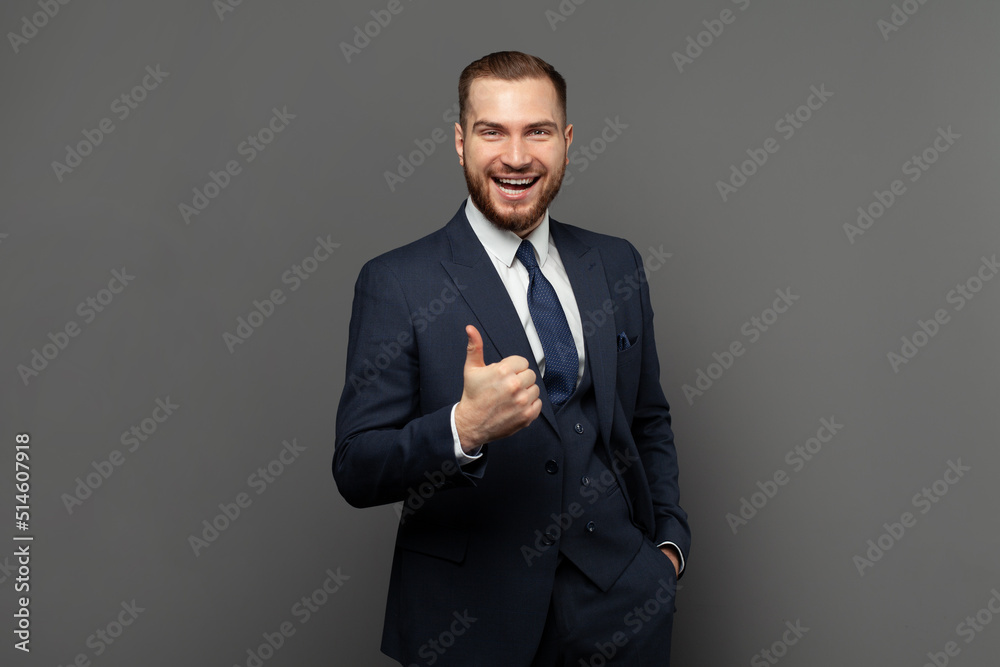 Happy handsome businessman wearing suit showing thumb up against grey studio wall background Happy handsome businessman wearing suit showing thumb up against grey studio wall background