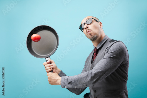 A funny chef man in suit holding frying pan and juggling tomato isolated over blue wall background.