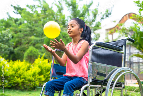 concept of happiness, freedom and enjoyment showing by Smiling alone girl kid with disability playing with ball while on wheelchair at park.