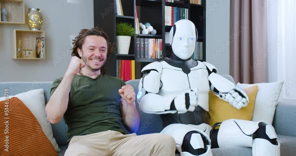Cheerful caucasian young man with robotic cyborg watching sport game on TV while sitting on comfy sofa at home. Humanoid machine with smiling male celebrating victory on championship.