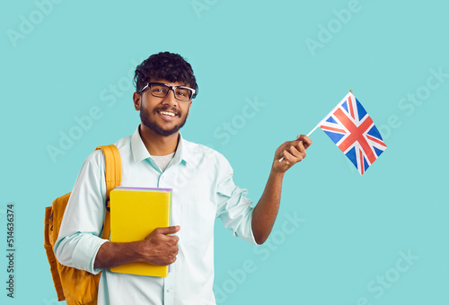 Studying in England. Portrait of happy Indian male university student with UK flag on light blue background. Ethnic smart guy with backpack and notebooks waving England flag looking at camera.