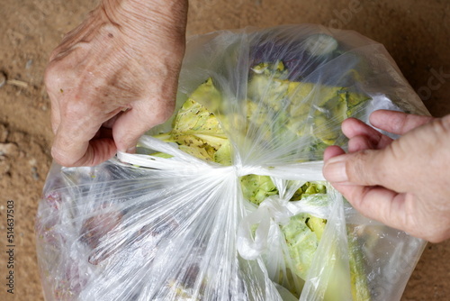 An old woman's hand is tying up a plastic bag with vegetable scraps and rambutan peels before discarding