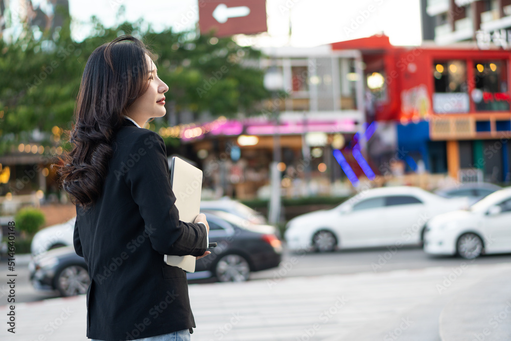 A young Asian businesswoman holding a laptop outdoors is walking into a company.