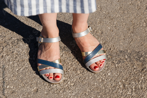 A close-up photo of women's feet in sandals with a t-shaped strap standing in the city in summer. Urban background. Feet in sandals close-up