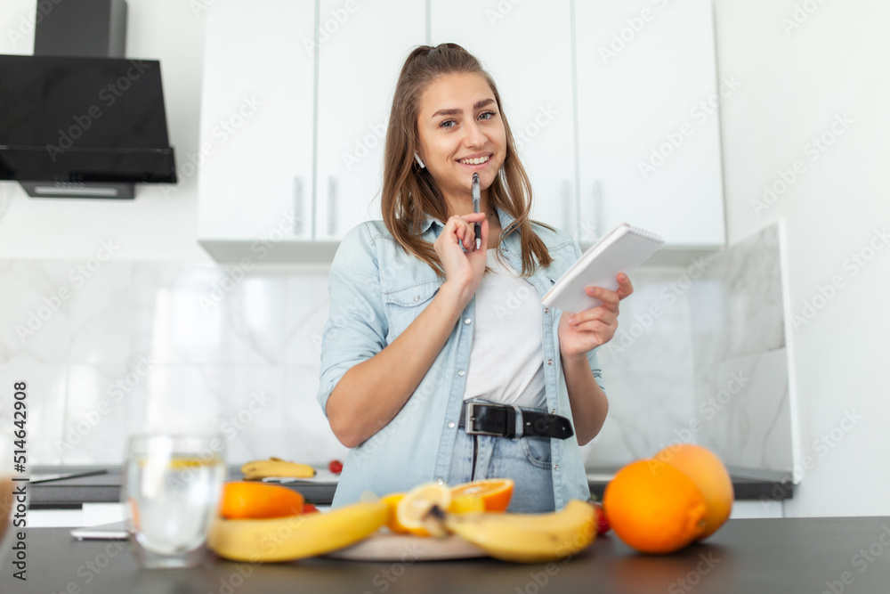 Young beautiful pensive woman with a notepad in the kitchen with fruits and vegetables. Recipe for cooking. Healthy food concept