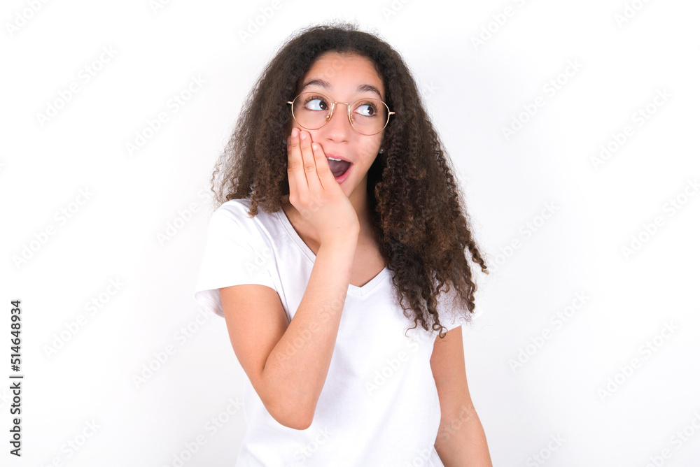 young beautiful girl with afro hairstyle wearing white t-shirt over white wall excited looking to the side hand on face. Advertisement and amazement concept.