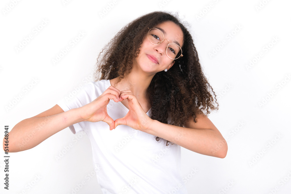 young beautiful girl with afro hairstyle wearing white t-shirt over white wall smiling in love showing heart symbol and shape with hands. Romantic concept. young beautiful girl with afro hairstyle wearing white t-shirt over white wall smiling in love showing heart symbol and shape with hands. Romantic concept.