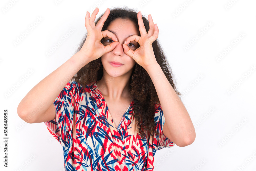 Playful excited young beautiful girl with afro hairstyle wearing flowered dress showing Ok sign with both hands on eyes, pretending to wear spectacles. Playful excited young beautiful girl with afro hairstyle wearing flowered dress showing Ok sign with both hands on eyes, pretending to wear spectacles.