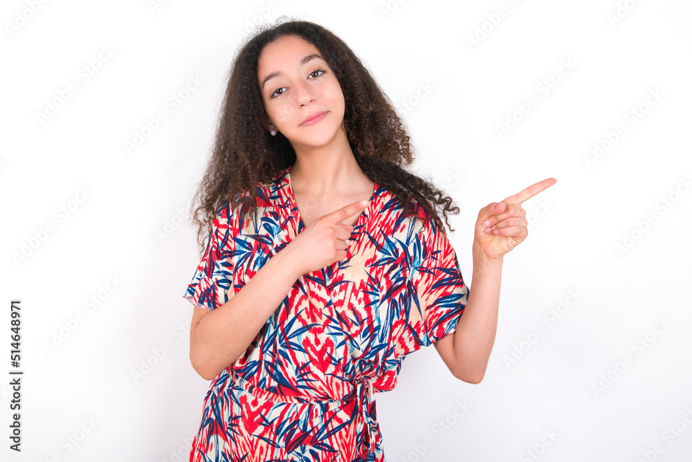 Positive young beautiful girl with afro hairstyle wearing flowered dress with beaming smile pointing with two fingers and looking on empty copy space. Advertisement concept. Positive young beautiful girl with afro hairstyle wearing flowered dress with beaming smile pointing with two fingers and looking on empty copy space. Advertisement concept.