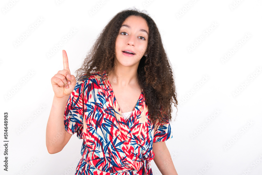 Pleasant looking young beautiful girl with afro hairstyle wearing flowered dress has clever expression, raises one finger, remembers herself not to forget tell important thing.