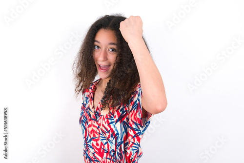 Overjoyed young beautiful girl with afro hairstyle wearing flowered dress glad to receive good news, clenching fist and making winning gesture. © Jihan