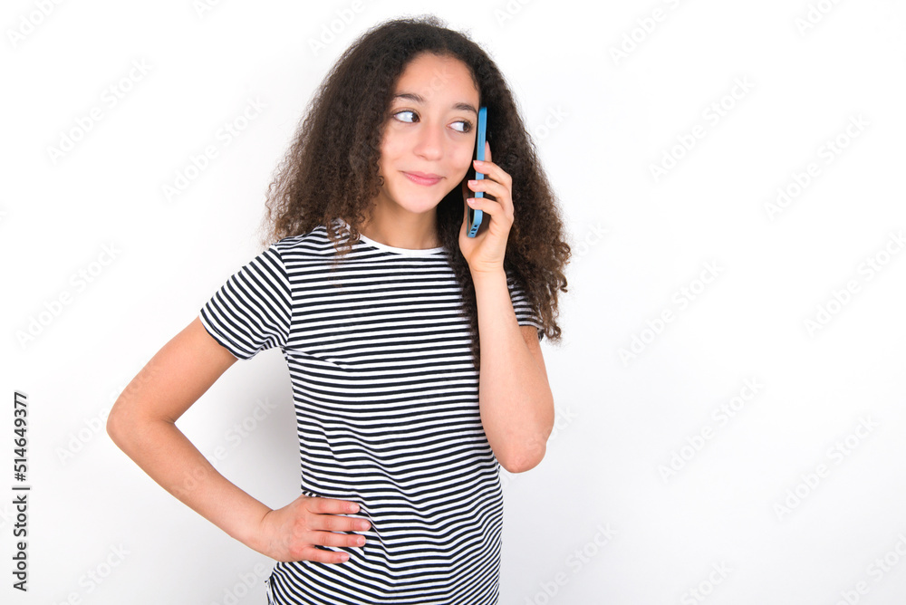Portrait of successful joyful young beautiful girl with afro hairstyle wearing striped t-shirt over white wall talking on mobile phone with friend. Lifestyle and communication concept Portrait of successful joyful young beautiful girl with afro hairstyle wearing striped t-shirt over white wall talking on mobile phone with friend. Lifestyle and communication concept
