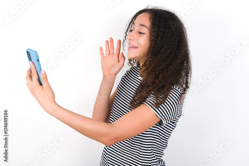 Portrait of happy friendly young beautiful girl with afro hairstyle wearing striped t-shirt over white wall taking selfie and waving hand, communicating on video call, online chatting.