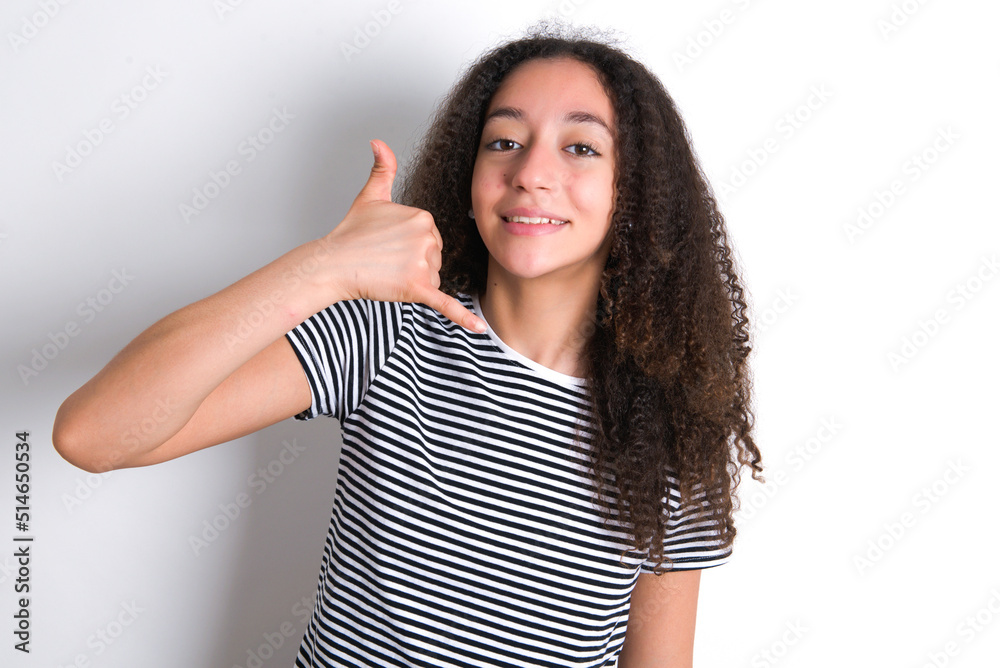 Φωτογραφία young beautiful girl with afro hairstyle wearing striped t-shirt over white wall makes phone gesture, says call me back again, has glad expression
