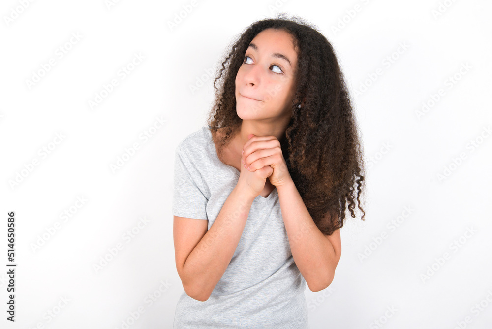Curious young beautiful girl with afro hairstyle wearing grey t-shirt over white wall keeps hands under chin bites lips and looks with interest aside. Curious young beautiful girl with afro hairstyle wearing grey t-shirt over white wall keeps hands under chin bites lips and looks with interest aside.