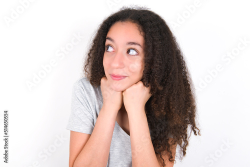 young beautiful girl with afro hairstyle wearing grey t-shirt over white wall holds hands under chin, glad to hear heartwarming words from stranger
