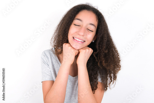 young beautiful girl with afro hairstyle wearing grey t-shirt over white wall grins joyfully, imagines something pleasant, copy space. Pleasant emotions concept.