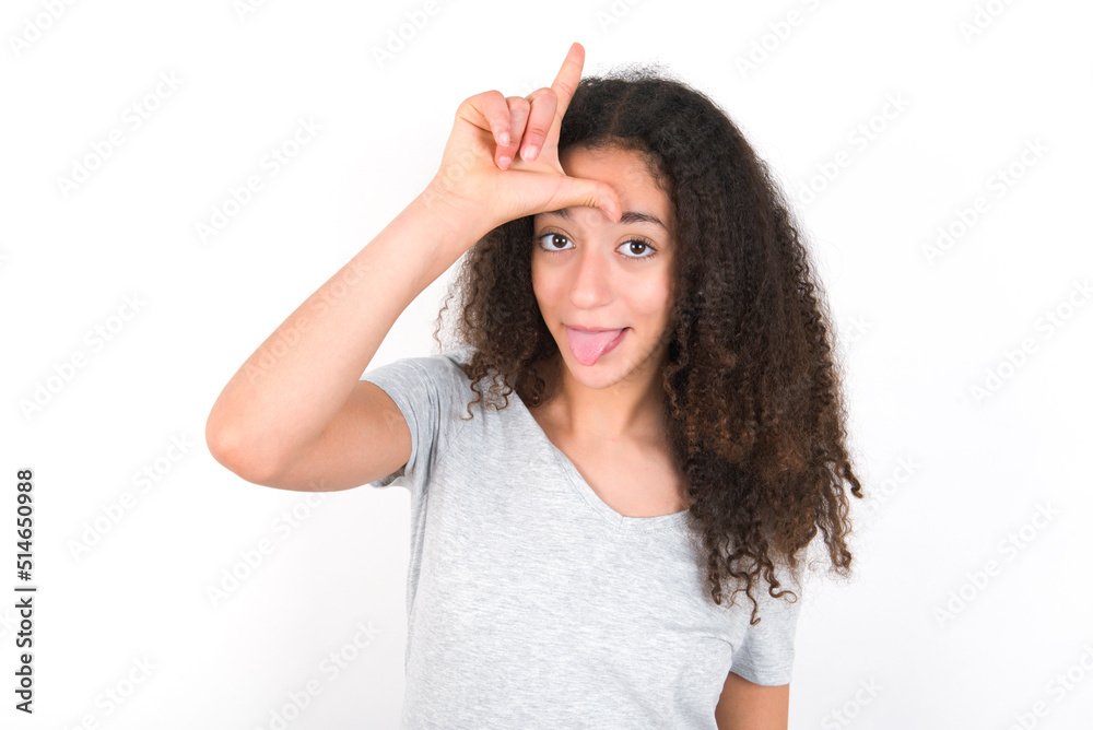 Funny young beautiful girl with afro hairstyle wearing grey t-shirt over white wall makes loser gesture mocking at someone sticks out tongue making grimace face. Funny young beautiful girl with afro hairstyle wearing grey t-shirt over white wall makes loser gesture mocking at someone sticks out tongue making grimace face.