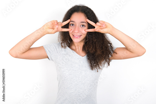 Cheerful positive young beautiful girl with afro hairstyle wearing grey t-shirt over white wall shows v-sign near eyes open mouth