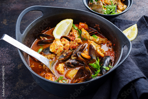 Modern style traditional Spanish seafood zarzuela de pescado with fish, king prawns and blue mussels served in red sauce as close-up in design pot