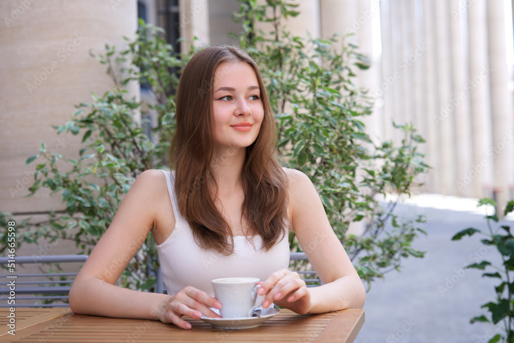 portrait of beautiful attractive happy smiling girl, young woman sitting outdoors on terrace in cafe on table drinking beverage tea or coffee at summer sunny day portrait of beautiful attractive happy smiling girl, young woman sitting outdoors on terrace in cafe on table drinking beverage tea or coffee at summer sunny day