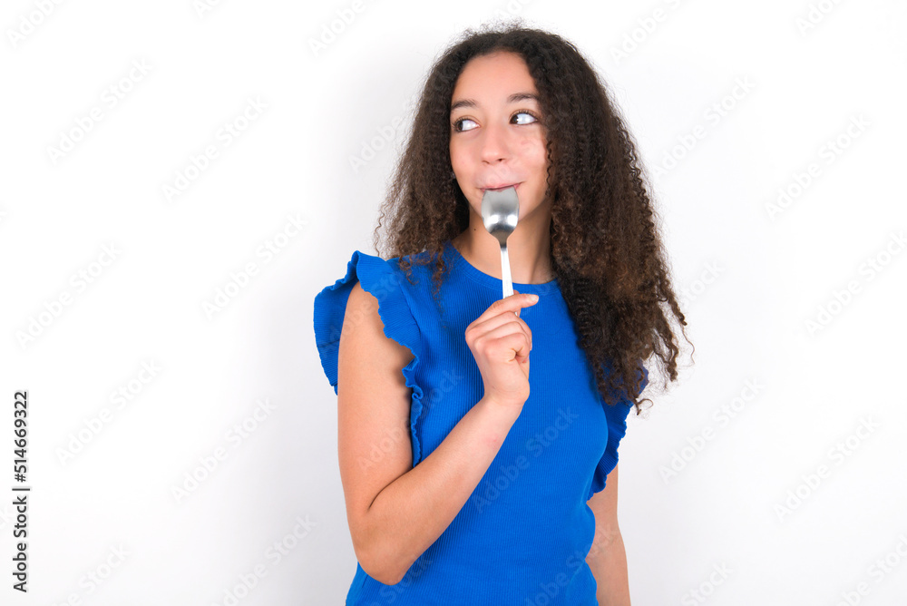 Very hungry Teenager girl with afro hairstyle wearing blue T-shirt over white wall holding spoon into mouth dream of tasty meal Very hungry Teenager girl with afro hairstyle wearing blue T-shirt over white wall holding spoon into mouth dream of tasty meal