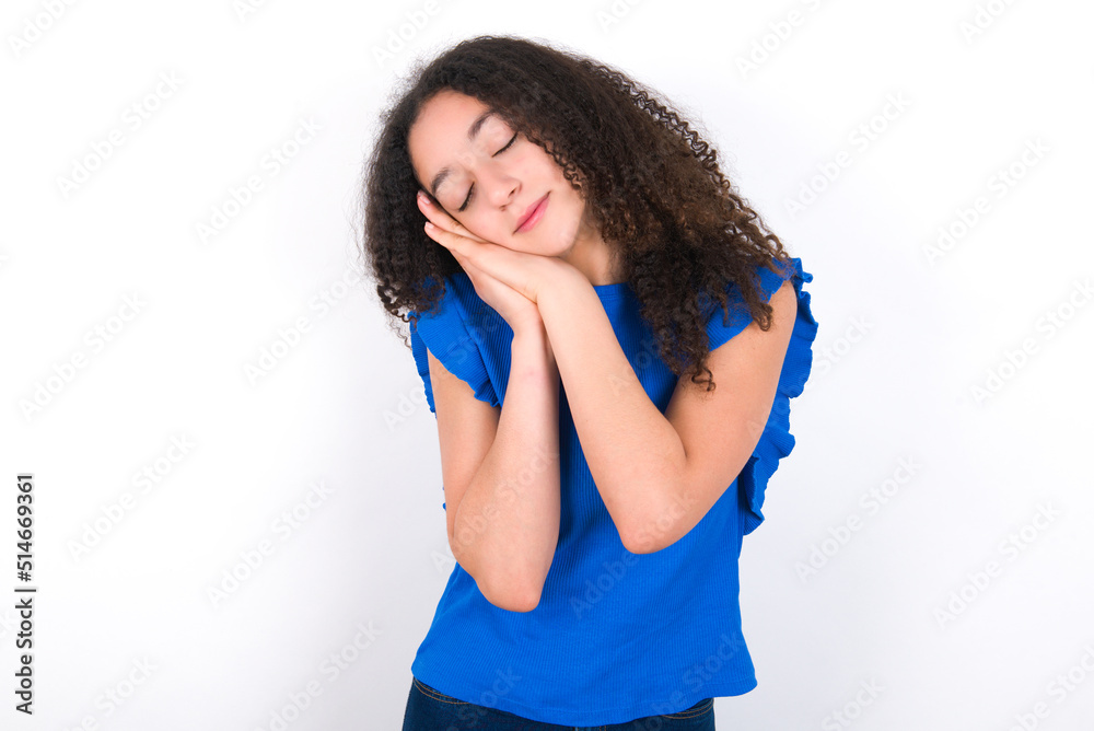 Teenager girl with afro hairstyle wearing blue T-shirt over white wall  sleeping tired dreaming and posing with hands together while smiling with closed eyes.