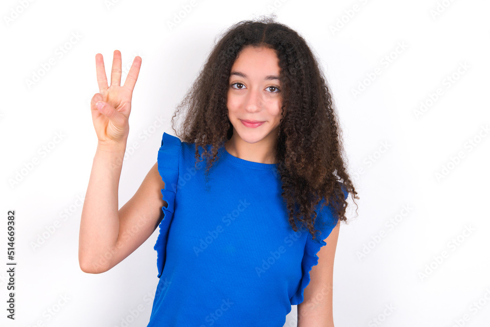Teenager girl with afro hairstyle wearing blue T-shirt over white wall showing and pointing up with fingers number three while smiling confident and happy. Teenager girl with afro hairstyle wearing blue T-shirt over white wall showing and pointing up with fingers number three while smiling confident and happy.